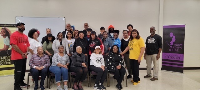 Group photo of diverse individuals gathered for a community event, showcasing unity and support, with a "Believe, Achieve, Receive" banner in the background.