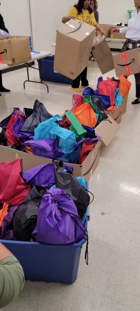 Colorful drawstring bags fill blue bins, while volunteers sort supplies into boxes in a community setting, highlighting a preparation event for distribution.