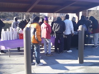 People gather around a table with cups and refreshments at an outdoor event, highlighting community engagement and social interaction in a park setting.