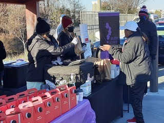 Volunteers distribute food and supplies at a community event, with tables set up for service and promotional materials on display.