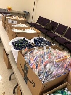 Boxes filled with medical supplies, including syringes and other equipment, are neatly arranged on tables in a community center.