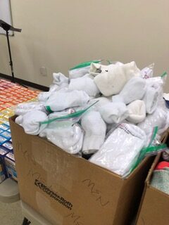 A cardboard box overflowing with neatly packed white socks in ziplock bags, indicating a donation or charity initiative for those in need.
