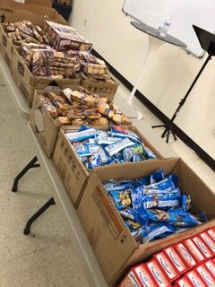 Boxes of assorted snacks, including granola bars and cookies, arranged on a table in a community space, highlighting food distribution efforts.