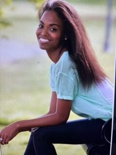 Smiling young woman in a light blue top sits casually outdoors, embodying a cheerful and relaxed atmosphere, relevant for lifestyle or fashion content.