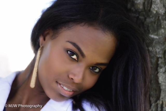 A woman with long, dark hair leans against a tree, smiling softly. She wears a white top and statement earrings, exuding confidence and grace.